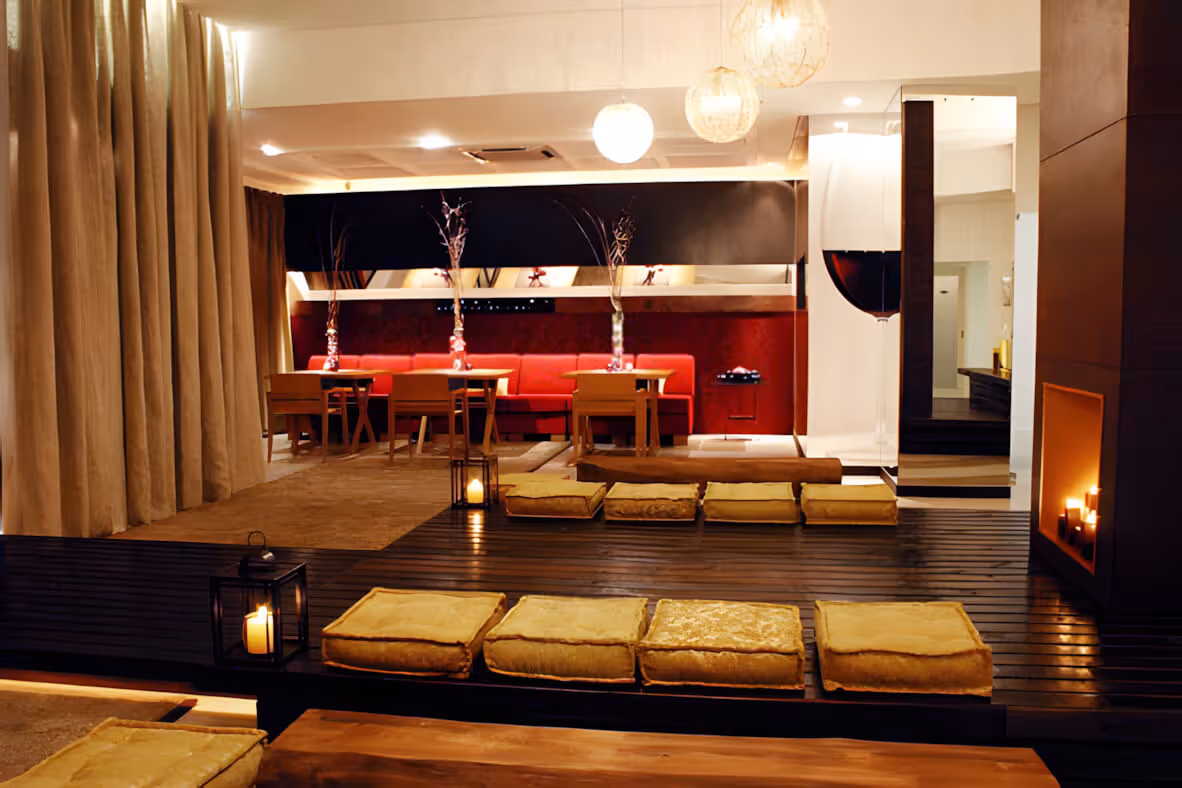 Cozy modern lounge with low seating cushions on dark wooden floor, small tables, red sofa, lantern candles, and warm ambient lighting.