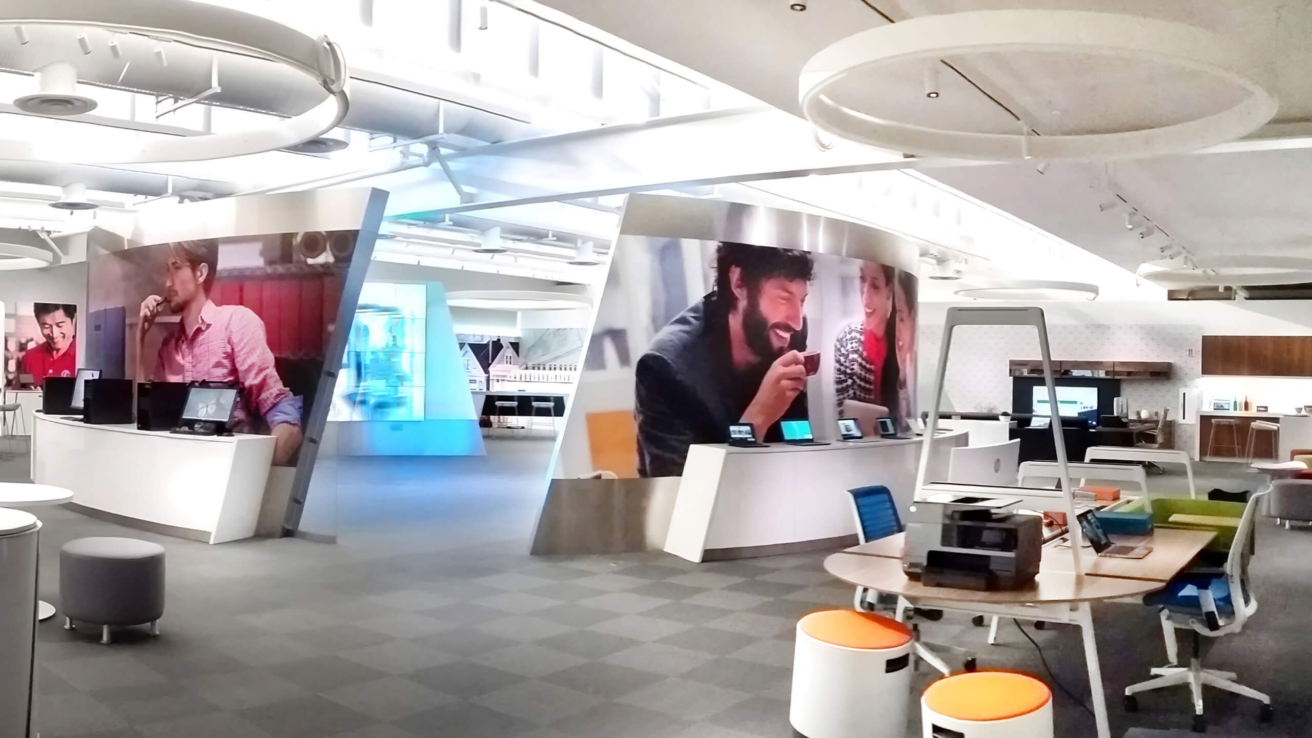 Modern office space with curved white desks, colorful seating, circular ceiling lights, and large wall screens displaying people.