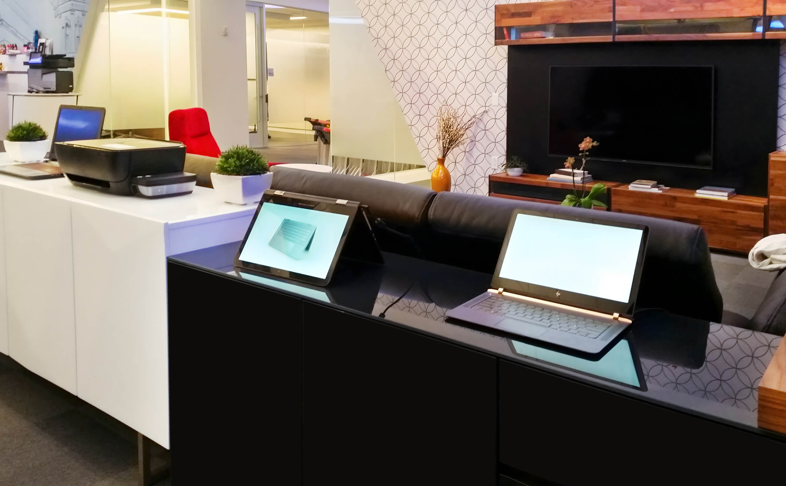 Modern office reception area with laptops on a black counter, a white cabinet with a printer, and a TV mounted on the wall.