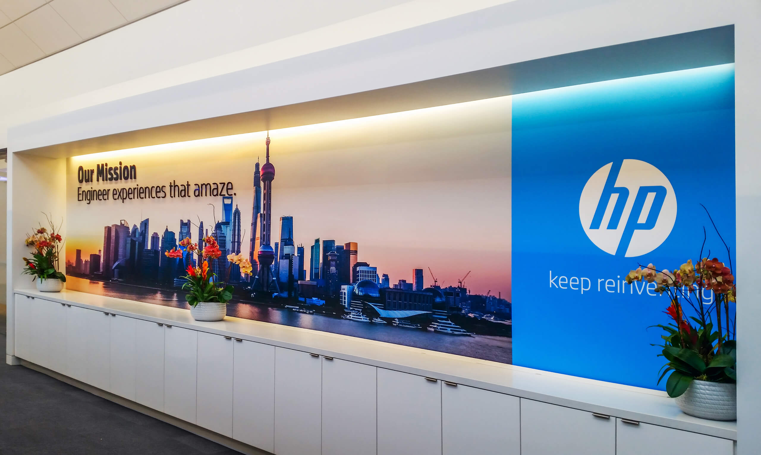 Office wall mural with a city skyline at sunset, text reading 'Our Mission Engineer experiences that amaze.', and the HP logo with 'keep reinventing.' slogan, flanked by flower arrangements.