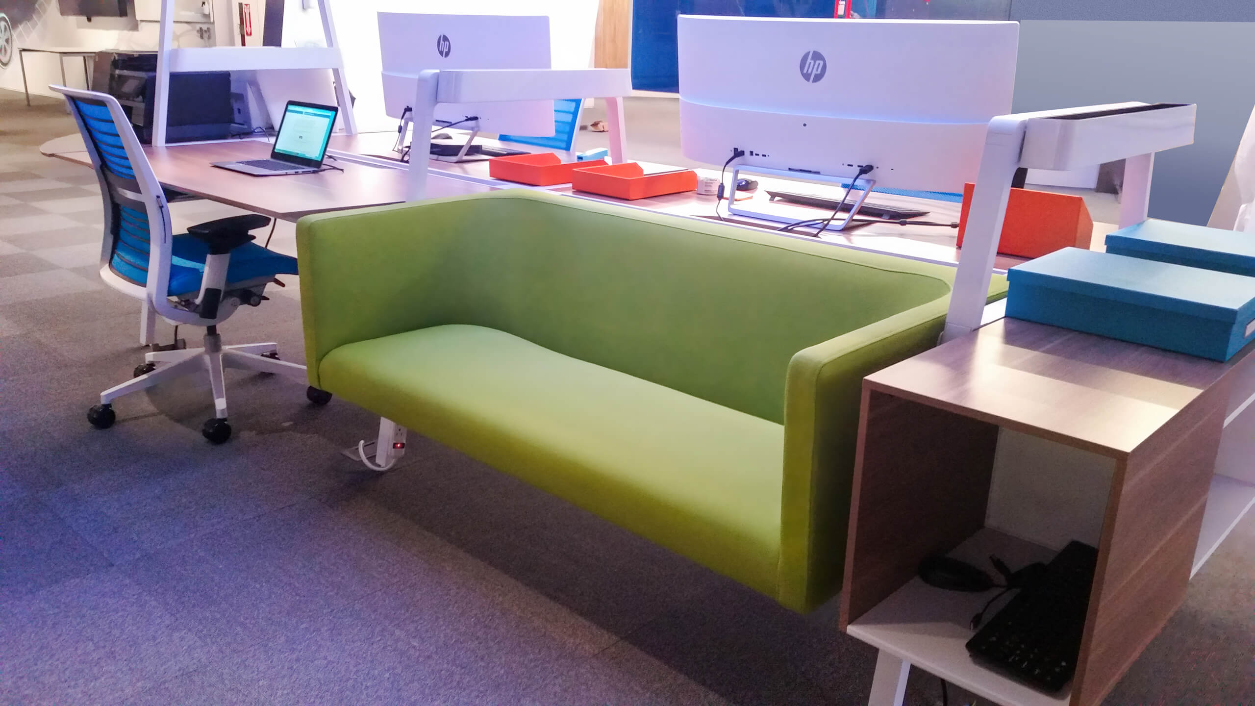 Modern office workspace with a green sofa attached to a desk, two HP monitors, a blue office chair, and a wooden side shelf holding a keyboard and mouse.