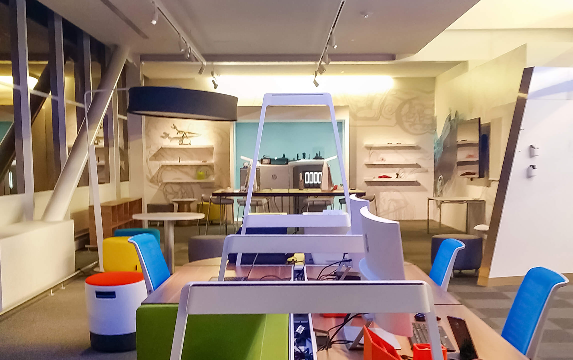 Modern office workspace with colorful chairs, white desks, computer monitors, and shelves on the far wall.