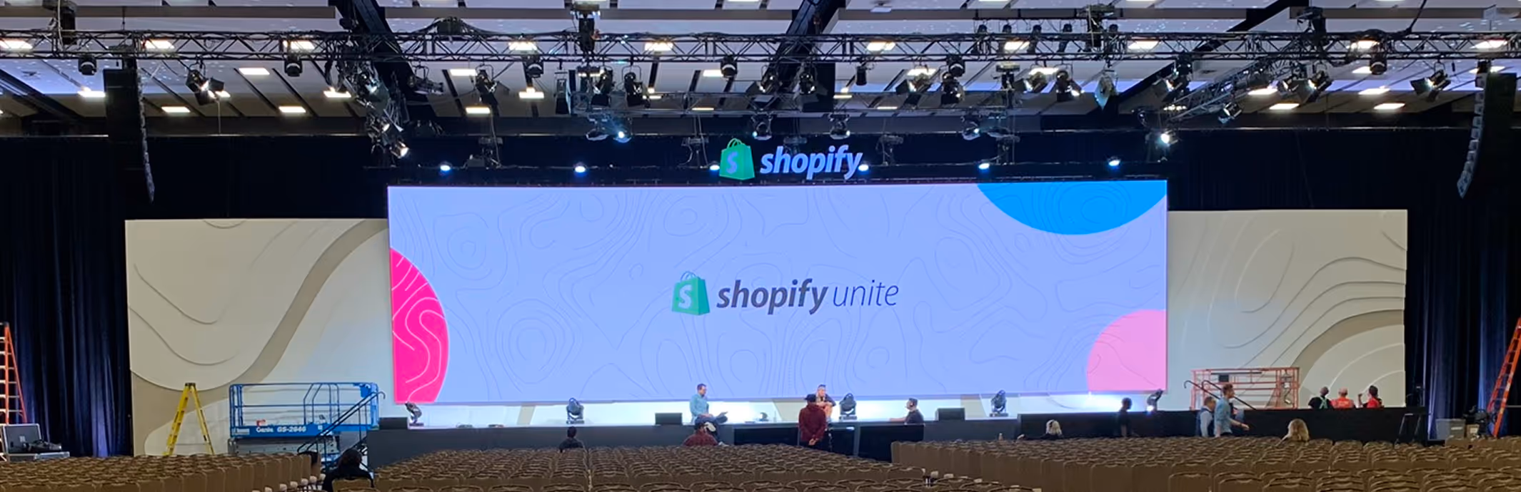 Large empty conference hall with rows of chairs facing a stage featuring a wide screen displaying the Shopify Unite logo.