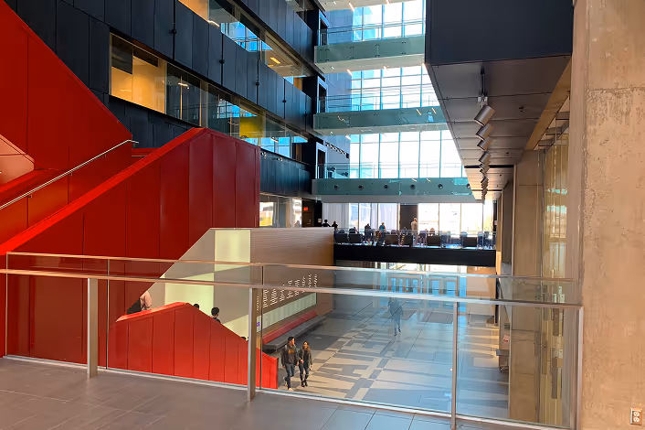 Modern indoor atrium with large windows, red staircases, glass railings, and people walking below.