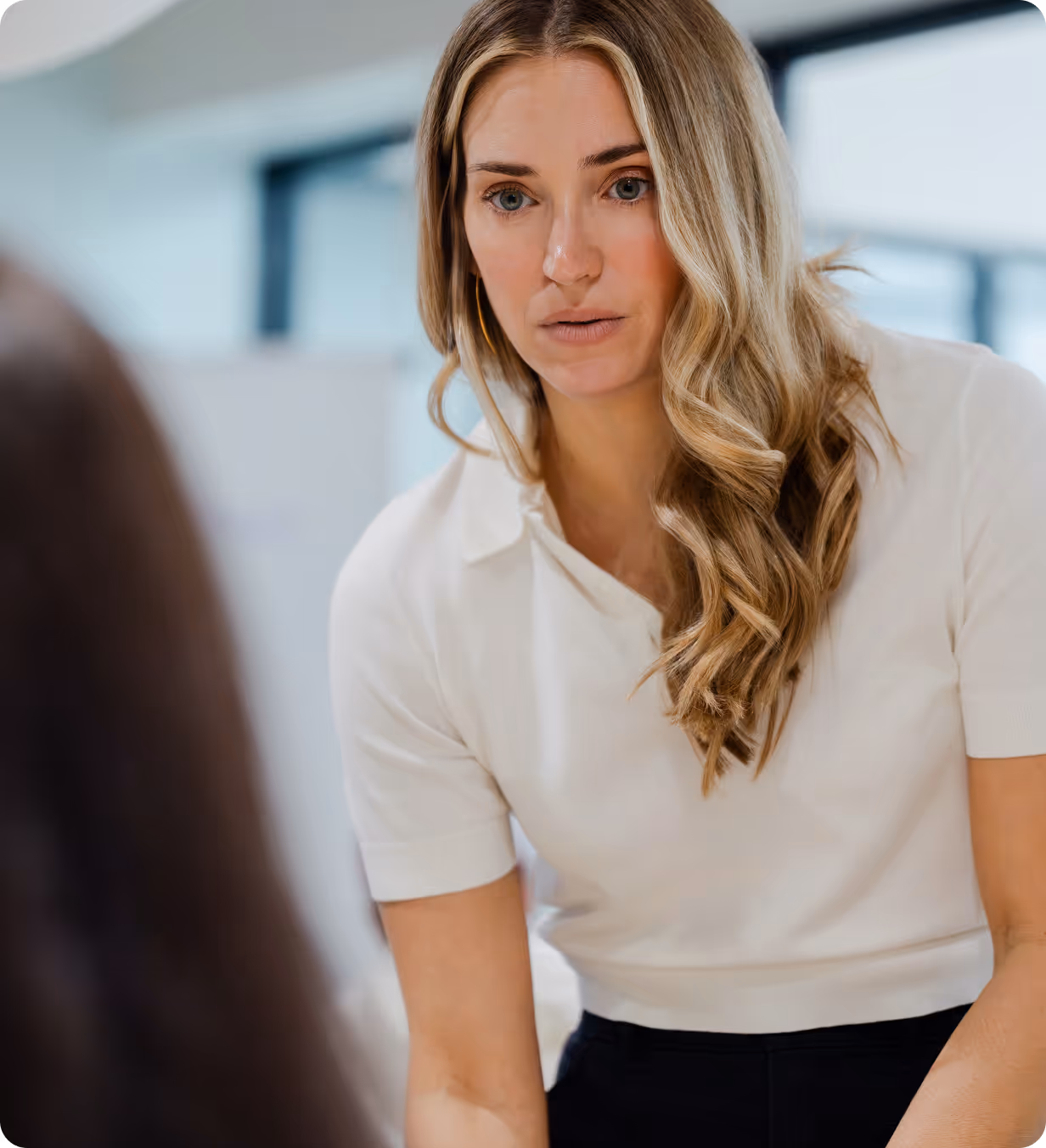 Focused woman with long blonde hair leaning forward, wearing a white shirt and gold hoop earrings in a modern office.