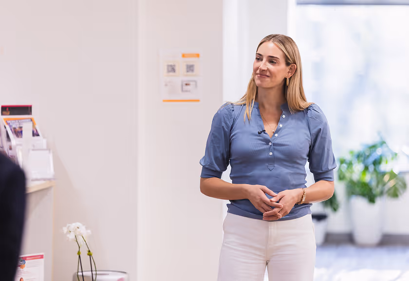 Smiling woman in blue blouse and white pants standing indoors in a bright room with plants in the background.