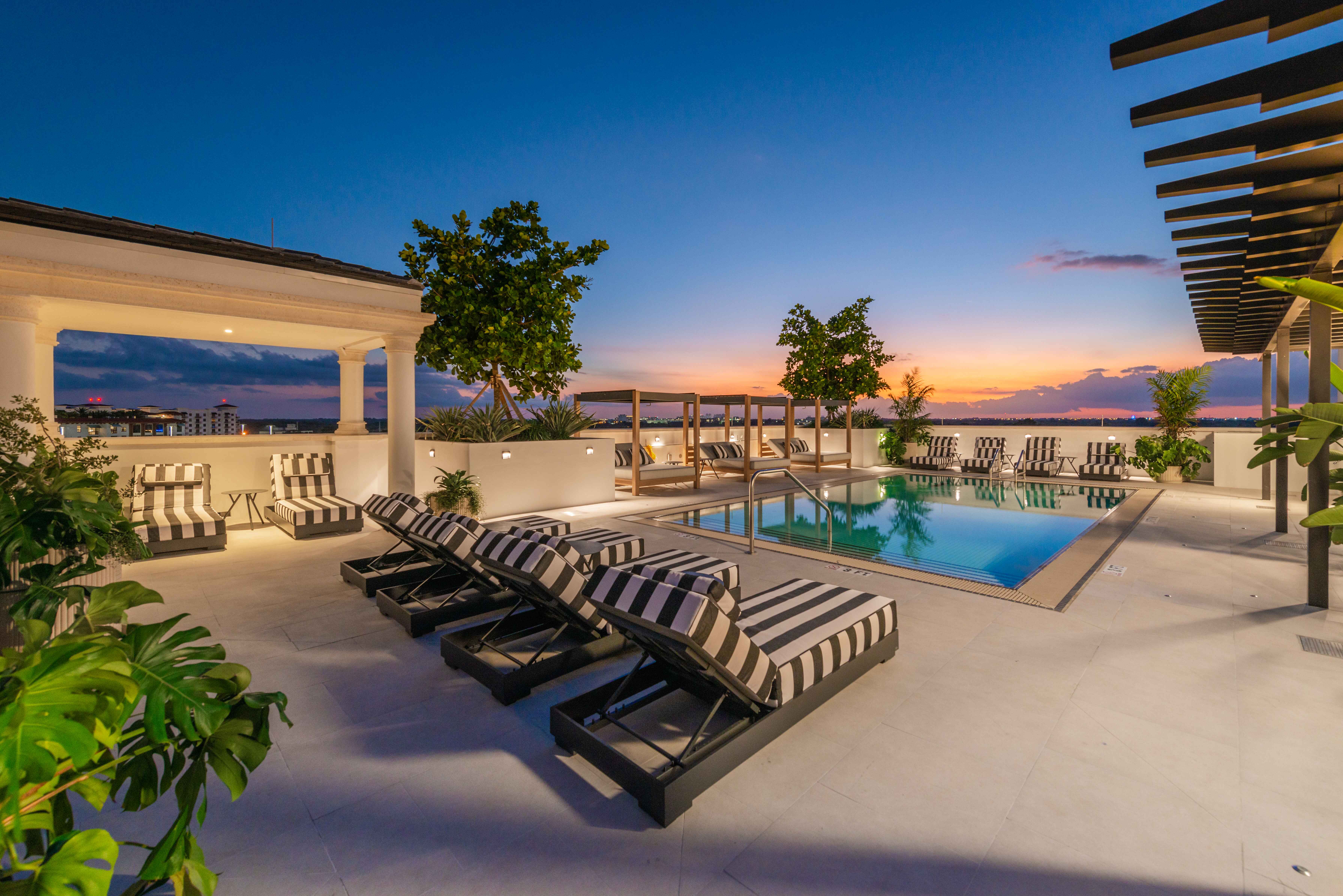 Our Rooftop pool area at sunset with striped lounge chairs, cabanas, plants, and city skyline views.