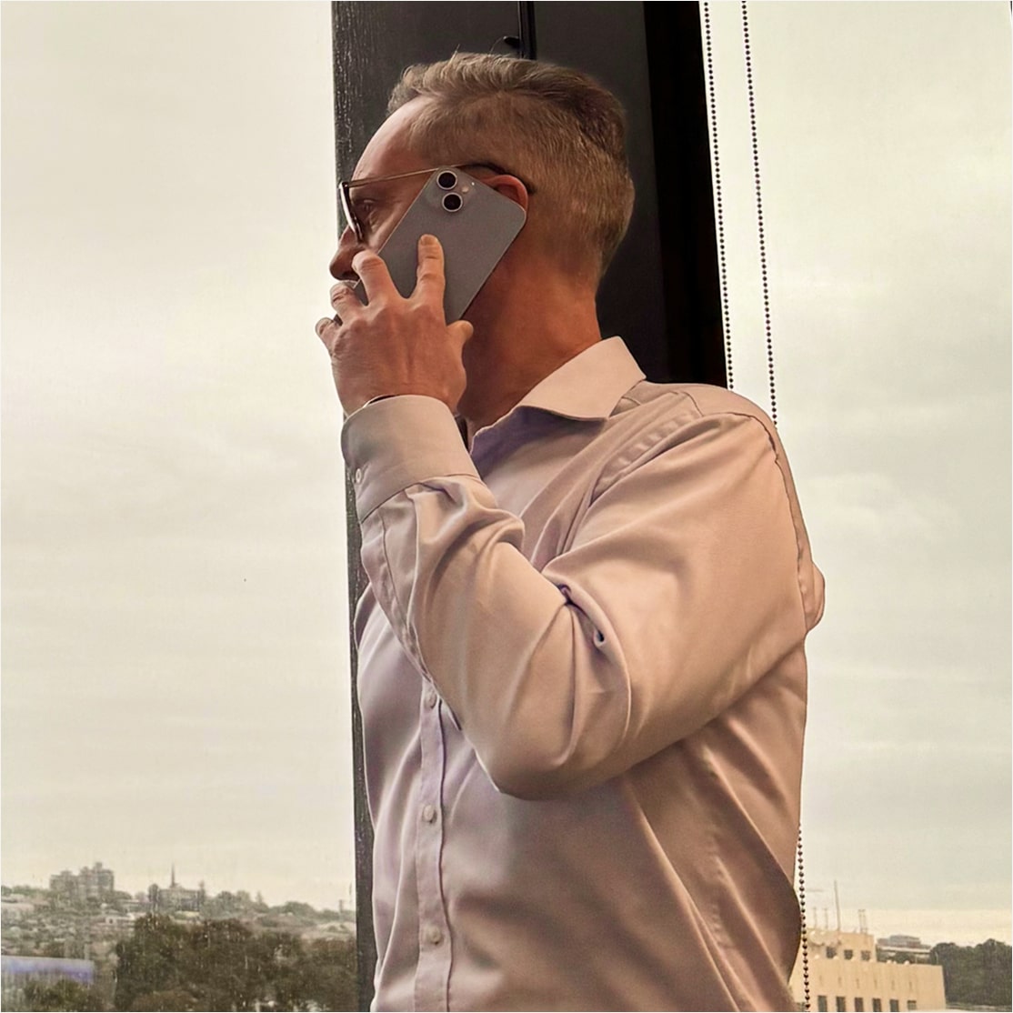 Man standing by a window on a phone call, wearing a light dress shirt and glasses, overlooking a cityscape on an overcast day.