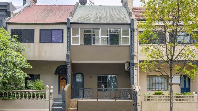Row of terrace houses in Sydney, with one central property featuring a grey facade, enclosed balcony, and decorative iron railings, framed by neighbouring homes and a tree in the foreground.
