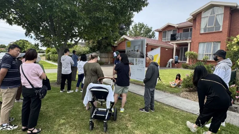 Group of potential homebuyers gathered on a suburban lawn outside a brick house, waiting for a property auction to begin. Some people stand near the auction sign while others sit or chat under a tree.