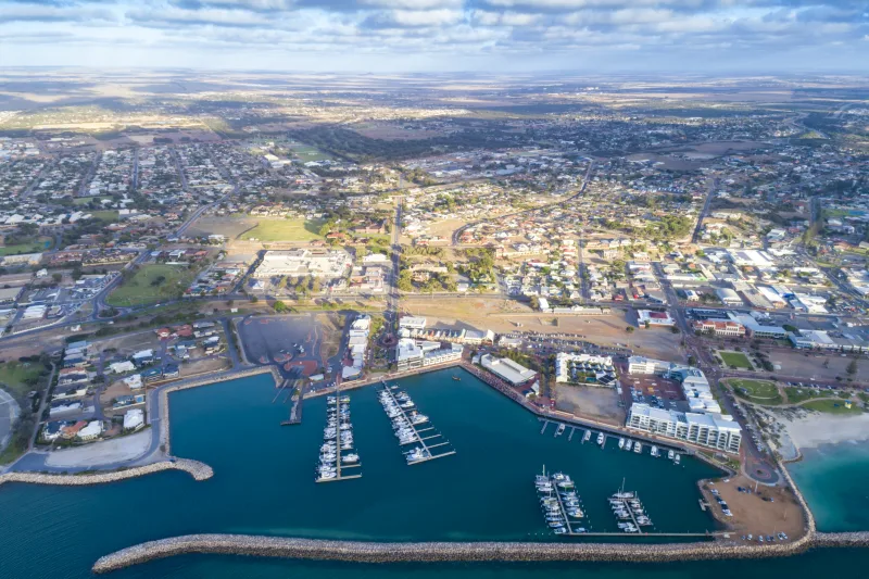 Aerial view of a coastal Australian city with a marina, boats docked in turquoise water, and surrounding residential and commercial areas extending inland under a partly cloudy sky.