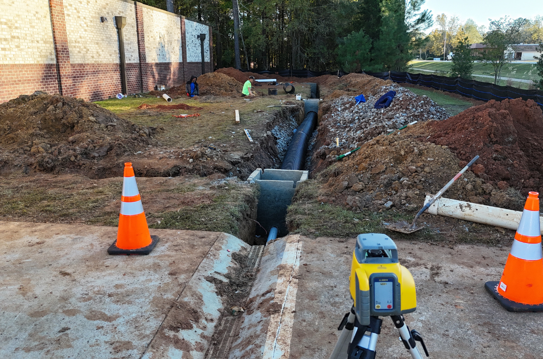 Construction site with a trench for pipe installation, orange safety cones, construction tools, and workers near a brick wall.