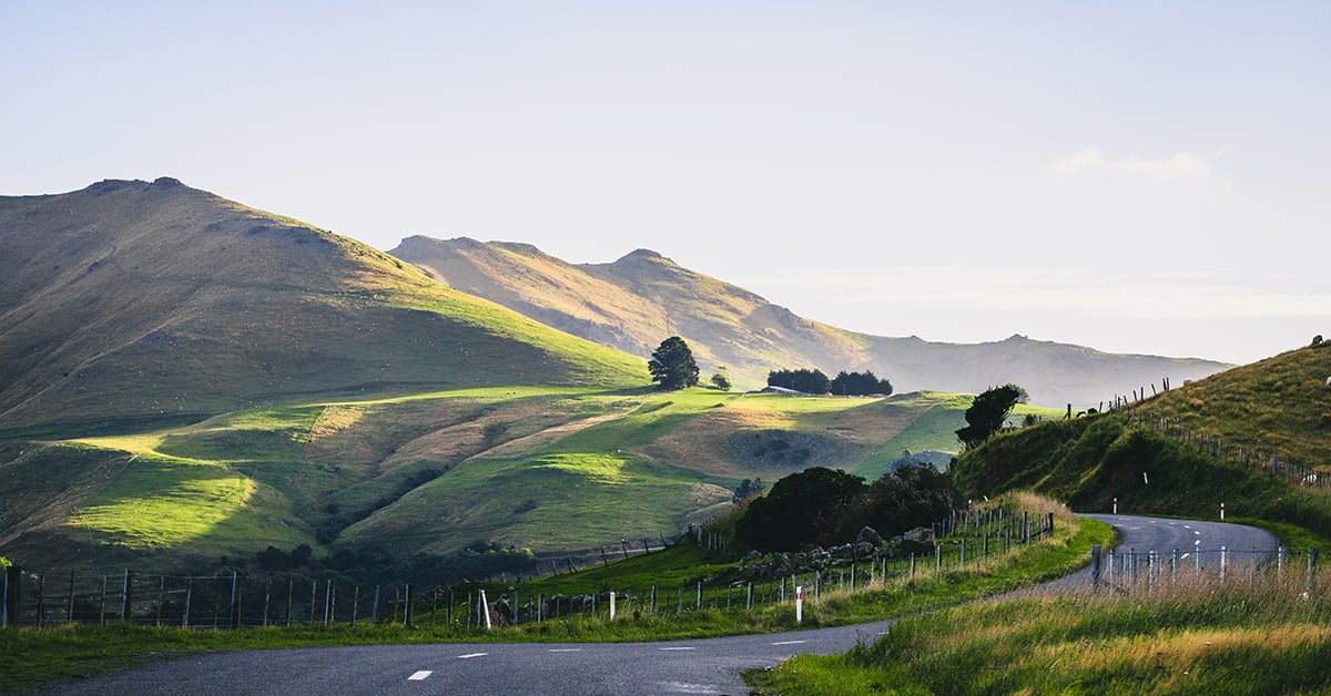 Winding road passing through green rolling hills and farmland under a clear sky.