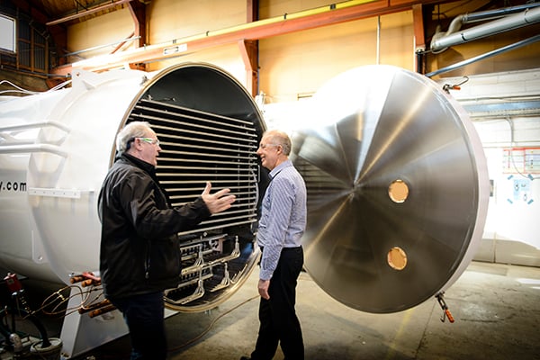 Two men talking beside a large industrial metal chamber with its door open inside a factory.