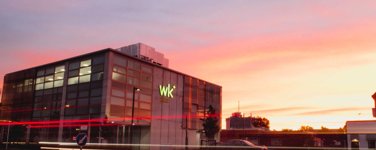 Modern office building with illuminated 'wk+' sign at sunset and streaks of red car lights.