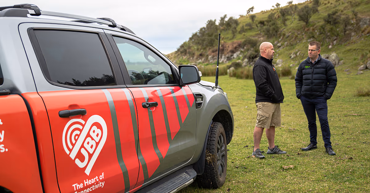 Two men standing on grass near a UBB truck with hills in the background.