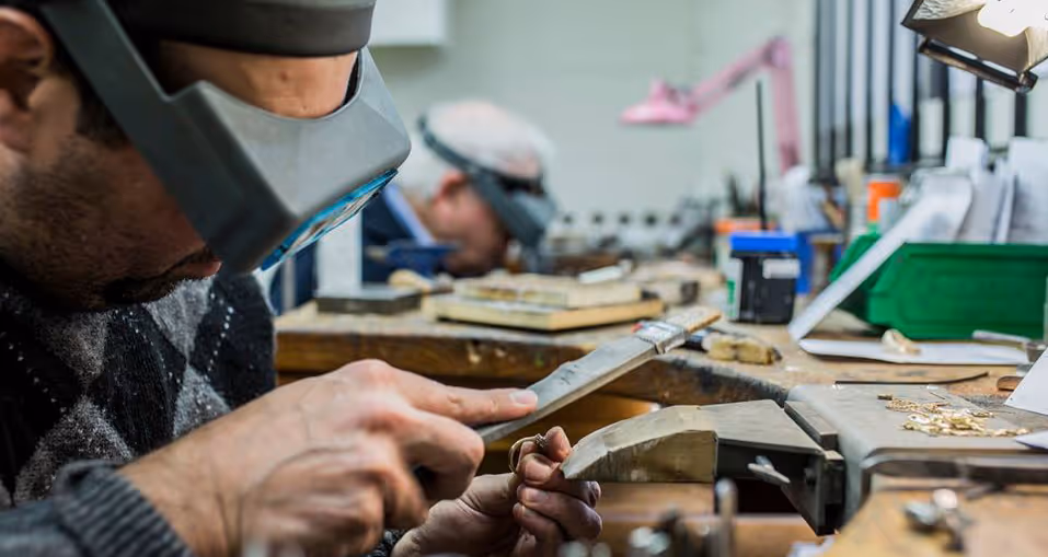 Two craftspeople wearing magnifying headsets working on detailed jewelry pieces at a cluttered workbench.