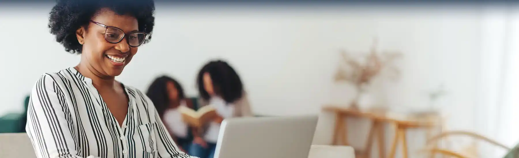 Smiling woman with glasses using laptop on a couch with two girls reading a book in the background.