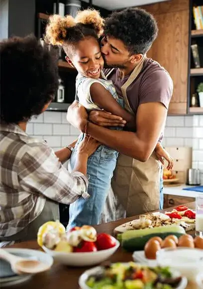 Father in an apron hugging and kissing his smiling daughter in a kitchen while the mother watches.