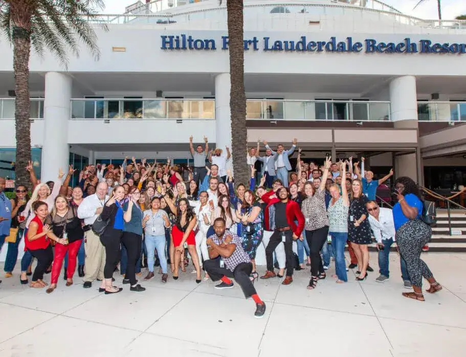 Large group of people posing cheerfully in front of the Hilton Fort Lauderdale Beach Resort, many with arms raised.