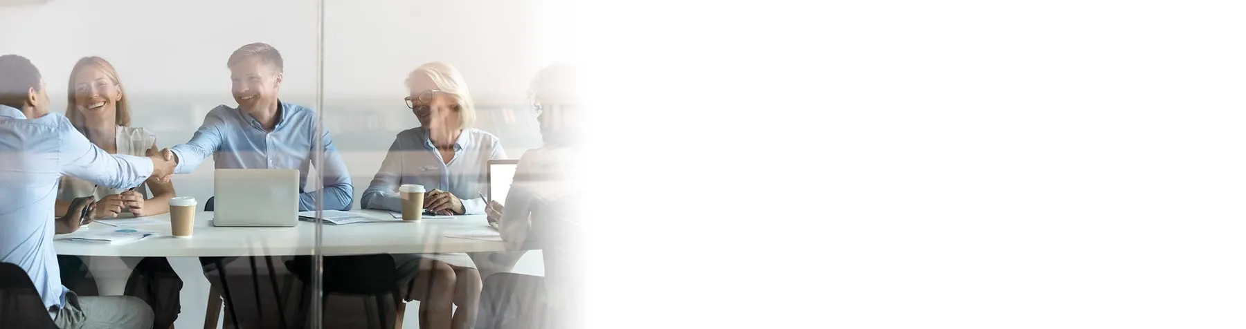 Five business professionals seated around a conference table, two men shaking hands, smiling during a meeting.