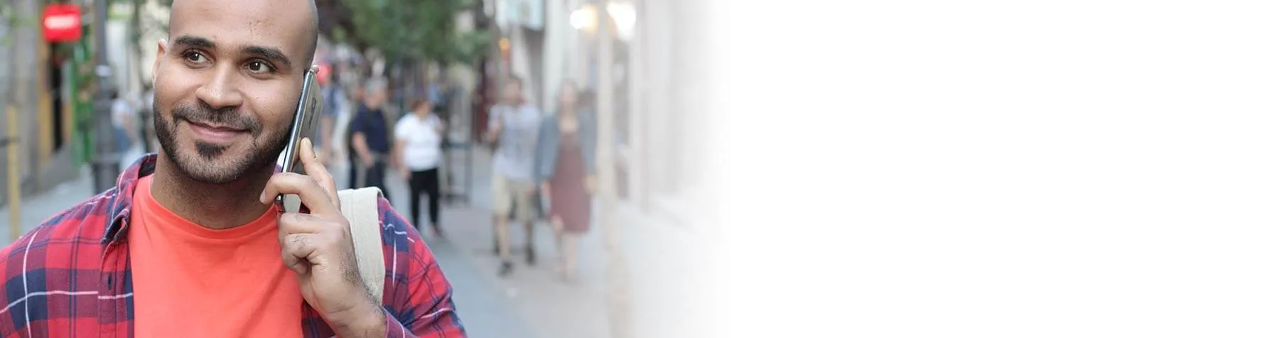 Smiling man wearing a red plaid shirt talking on a smartphone outdoors with blurred pedestrians in background.