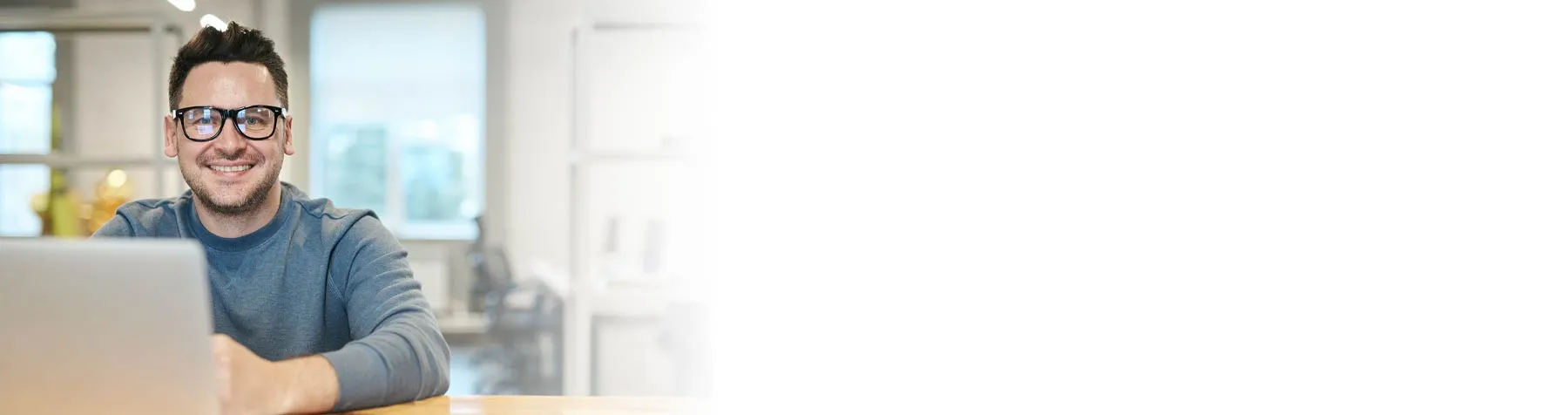 Smiling man wearing glasses sitting at a desk with a laptop in a bright office.