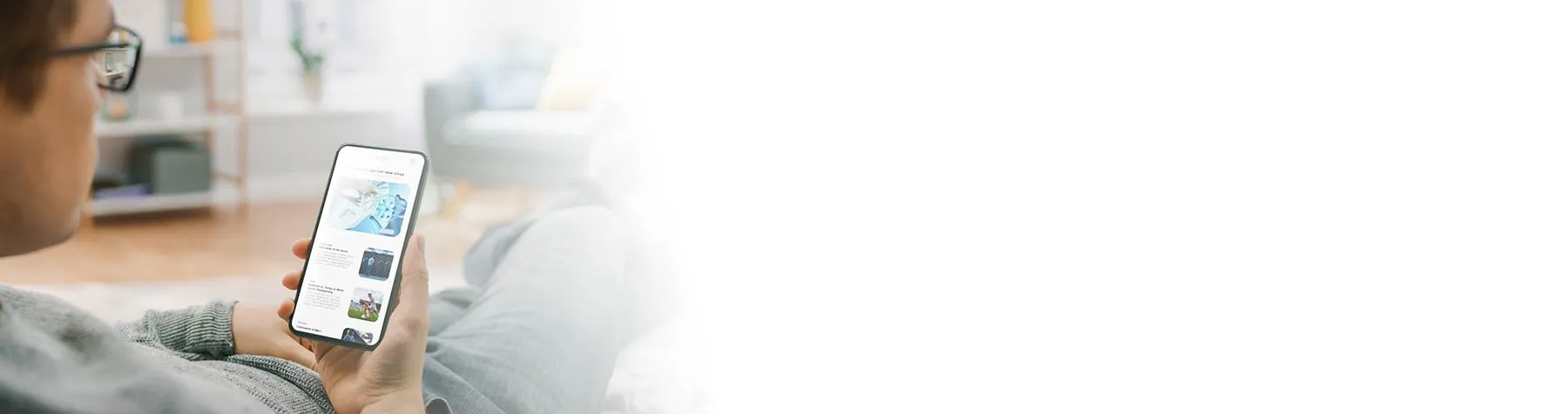 Person wearing glasses and a gray sweater sitting on a couch, holding and scrolling through a smartphone indoors.