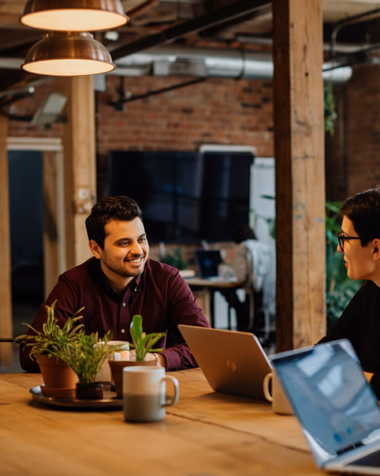 Two colleagues sitting at a wooden table with laptops, smiling and conversing in a modern office space.