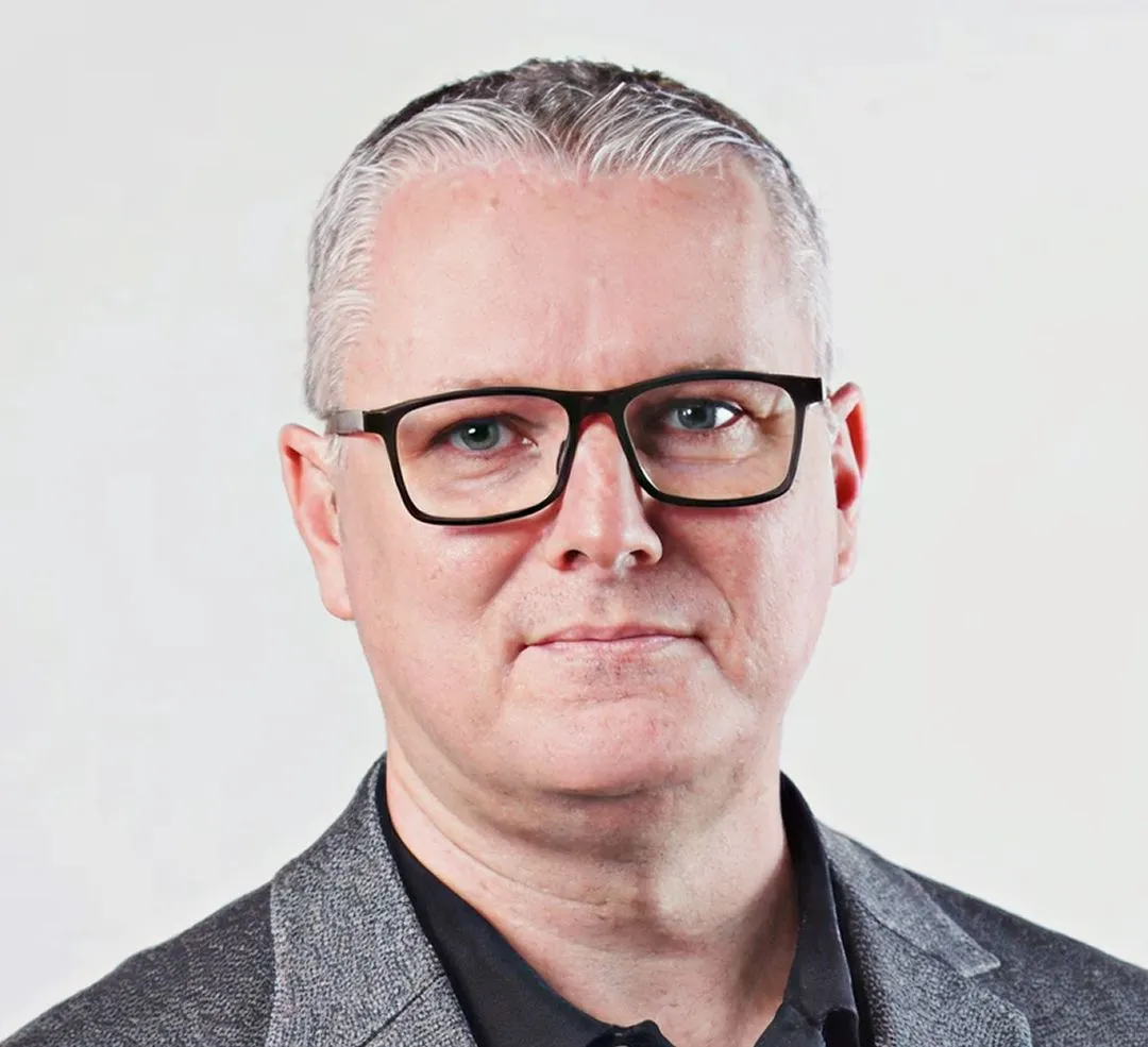 Middle-aged man with short gray hair, glasses, and a gray blazer over a black shirt, looking straight ahead.