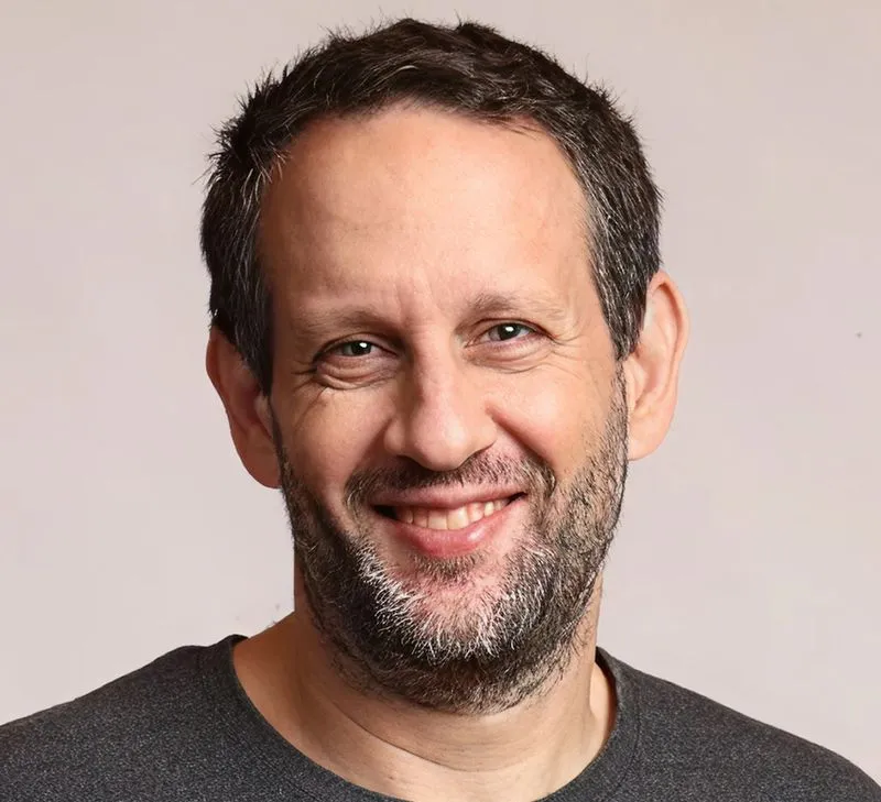 Smiling middle-aged man with short dark hair and a trimmed beard wearing a dark gray shirt against a plain light background.