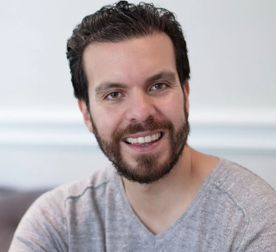 Smiling man with dark hair and beard wearing a light gray shirt, posing indoors.