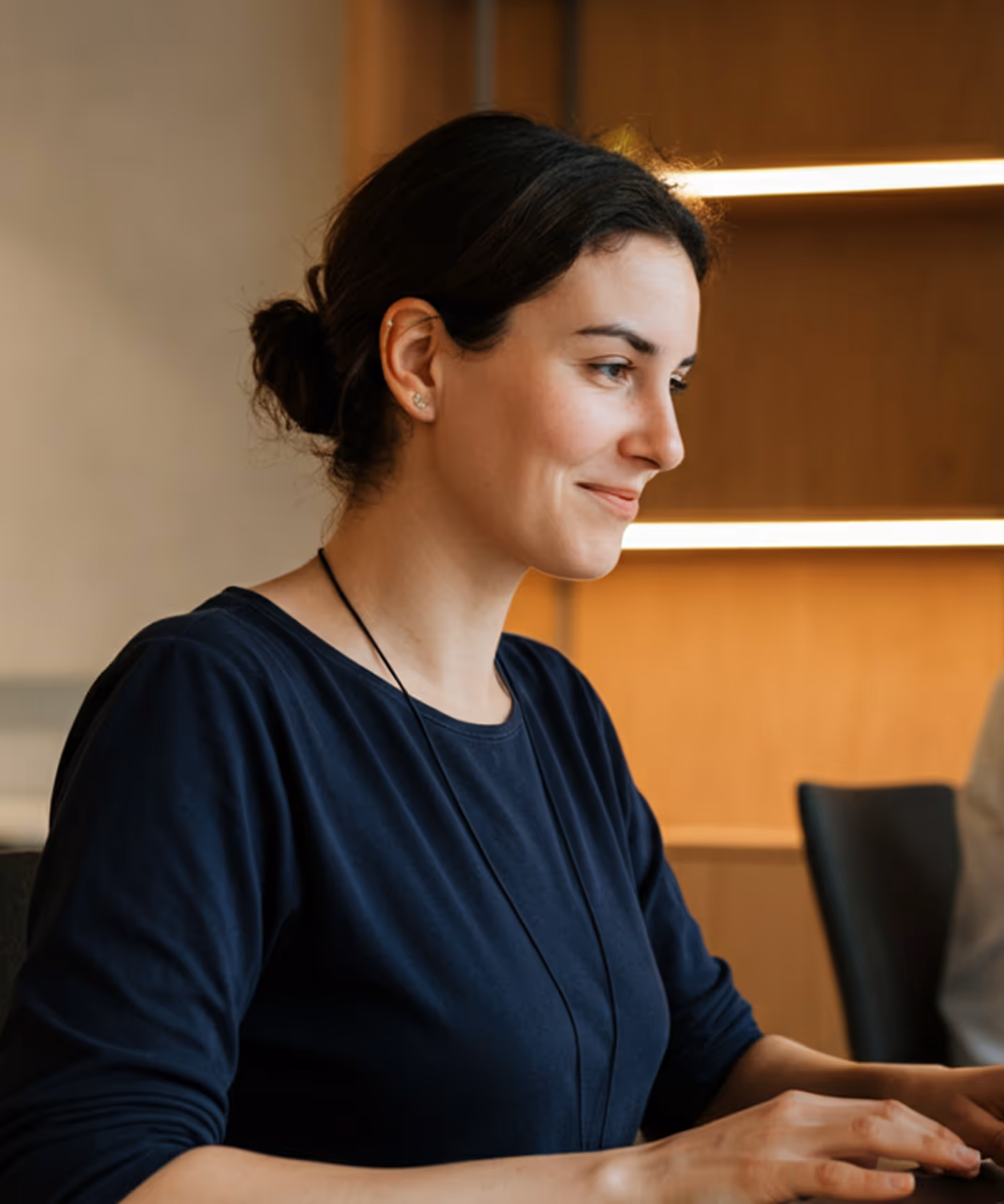 Young woman with dark hair in a bun, wearing a navy blue shirt, smiling while working at a computer.