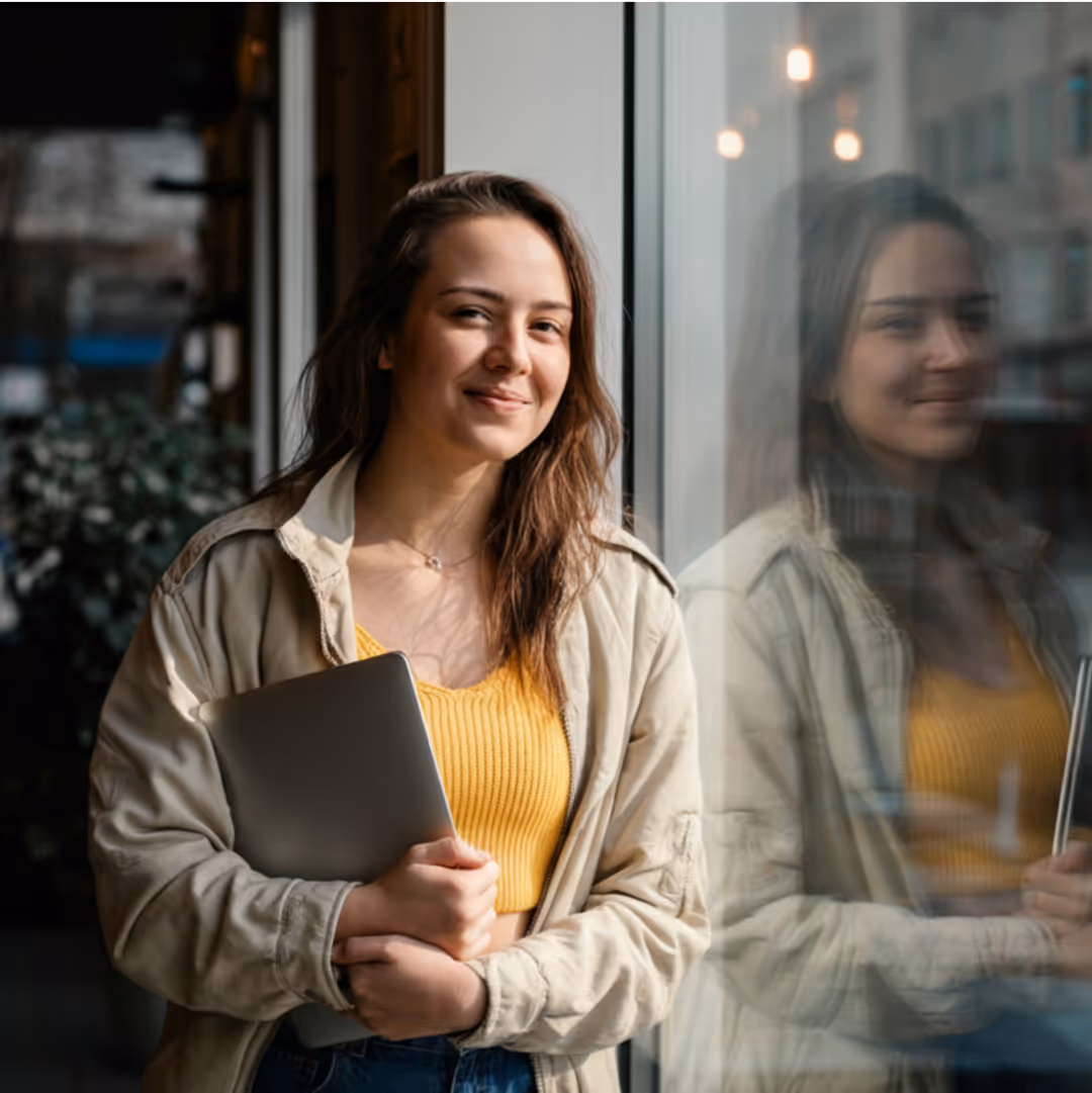 Young woman smiling and holding a laptop while standing next to a window with her reflection visible.
