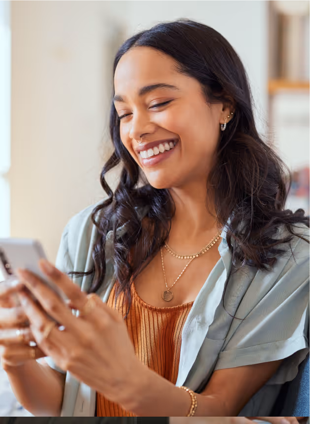 Smiling woman with long dark hair looking at a smartphone in her hands indoors.