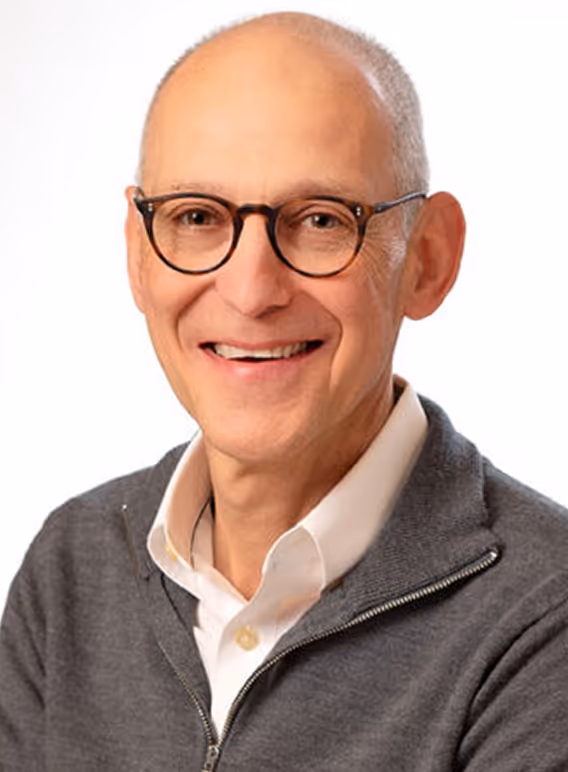 Smiling bald man wearing round glasses, white shirt, and grey zip-up sweater against a white background.