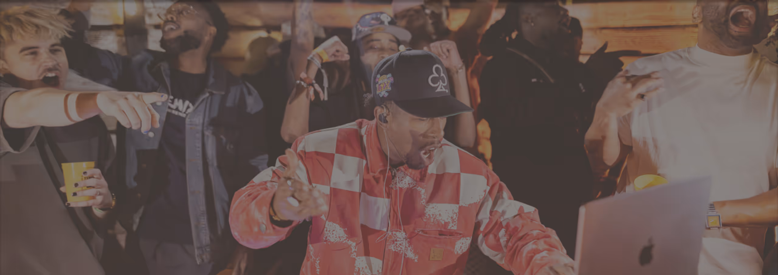 DJ wearing a checkered jacket and black cap performing at a lively party with cheering crowd around him.