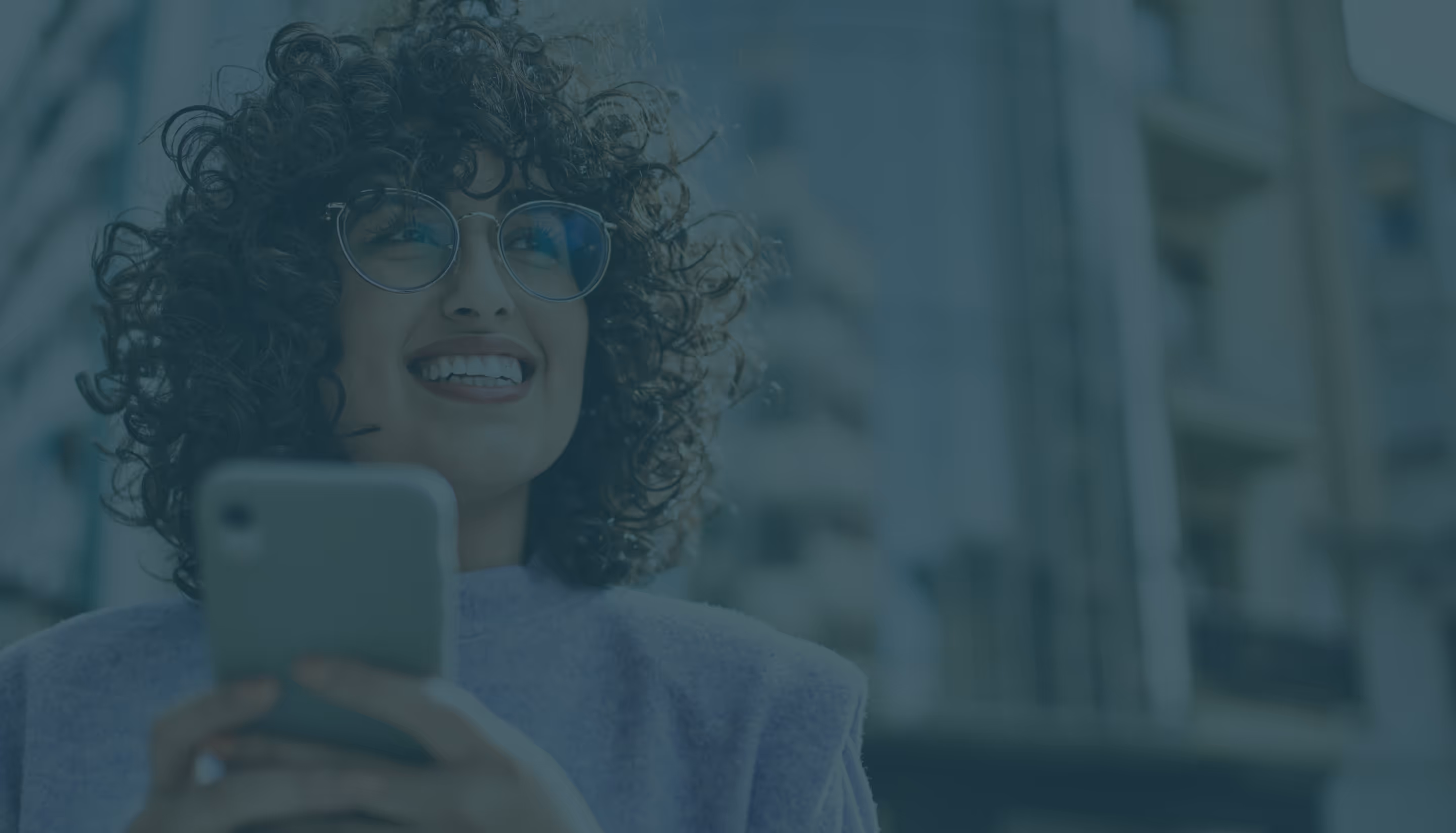Smiling woman with curly hair and glasses holding a smartphone outdoors in an urban setting.