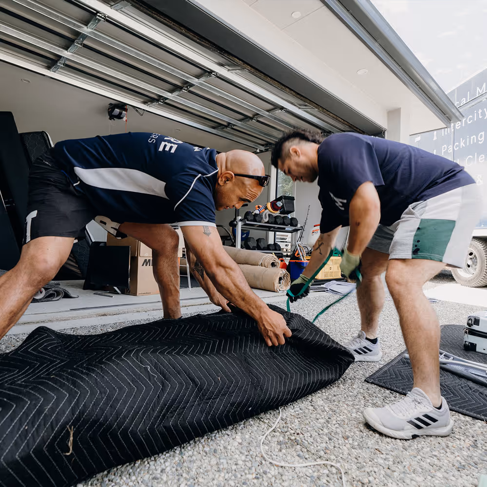 Two men in shorts and t-shirts working together to wrap a large item with a black moving blanket outside a garage.