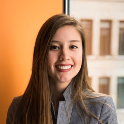 Portrait d'une jeune femme souriante aux longs cheveux bruns, portant un blazer gris, devant une fenêtre avec un mur orange en arrière-plan.