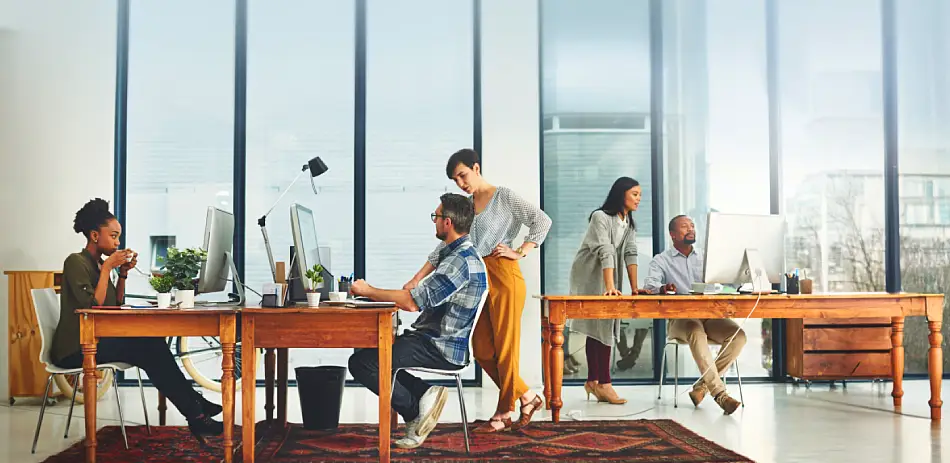 Five diverse coworkers in a modern office working at wooden desks with large windows in the background.