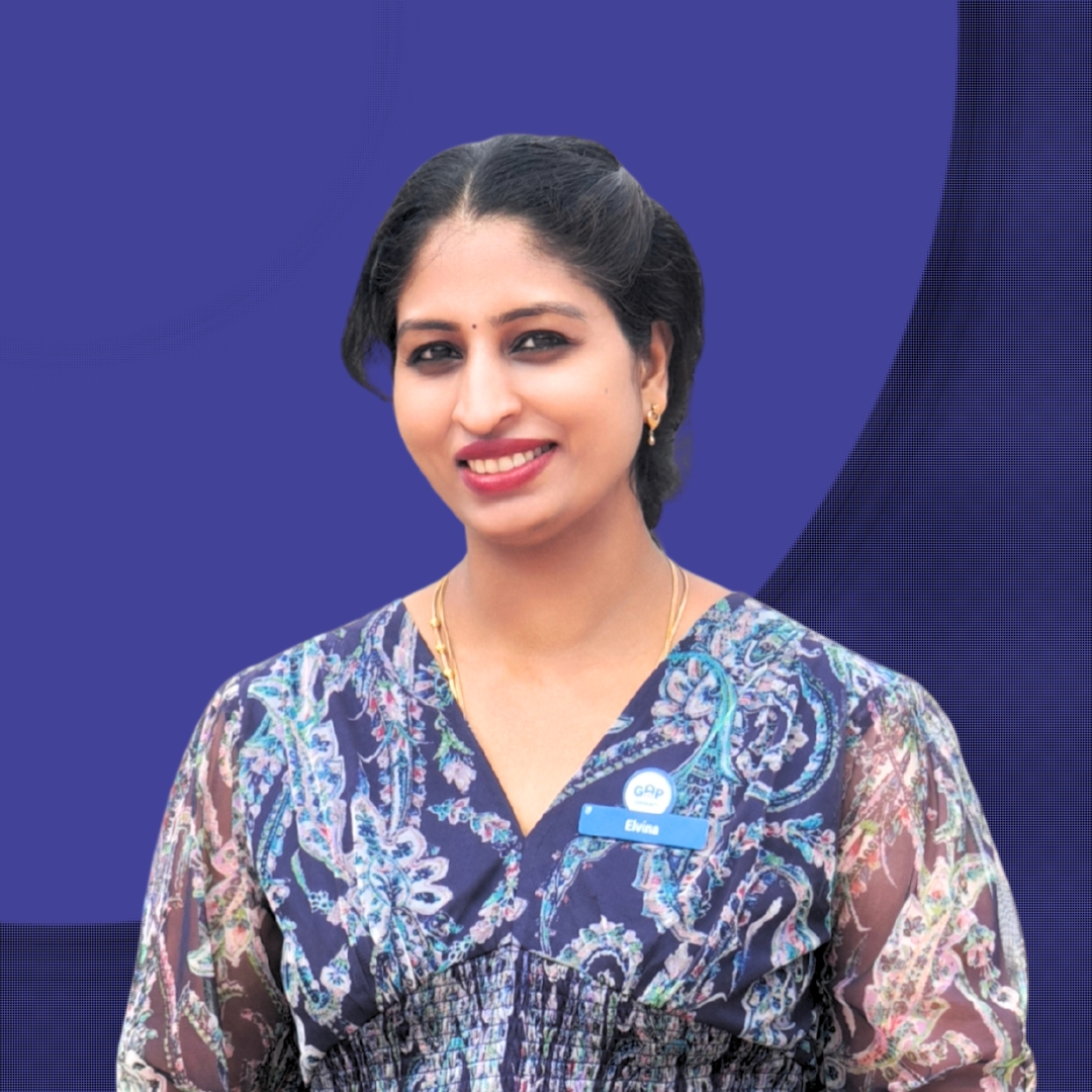 Smiling woman with black hair wearing a floral patterned blouse and a blue name tag labeled Elvina, against a blue background.