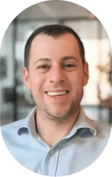 Portrait of a man with short dark hair, a beard, and black glasses wearing a light brown jacket and dark shirt.