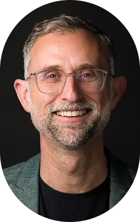 Young woman with medium-length dark hair wearing black glasses and silver earrings, smiling slightly against a plain background.