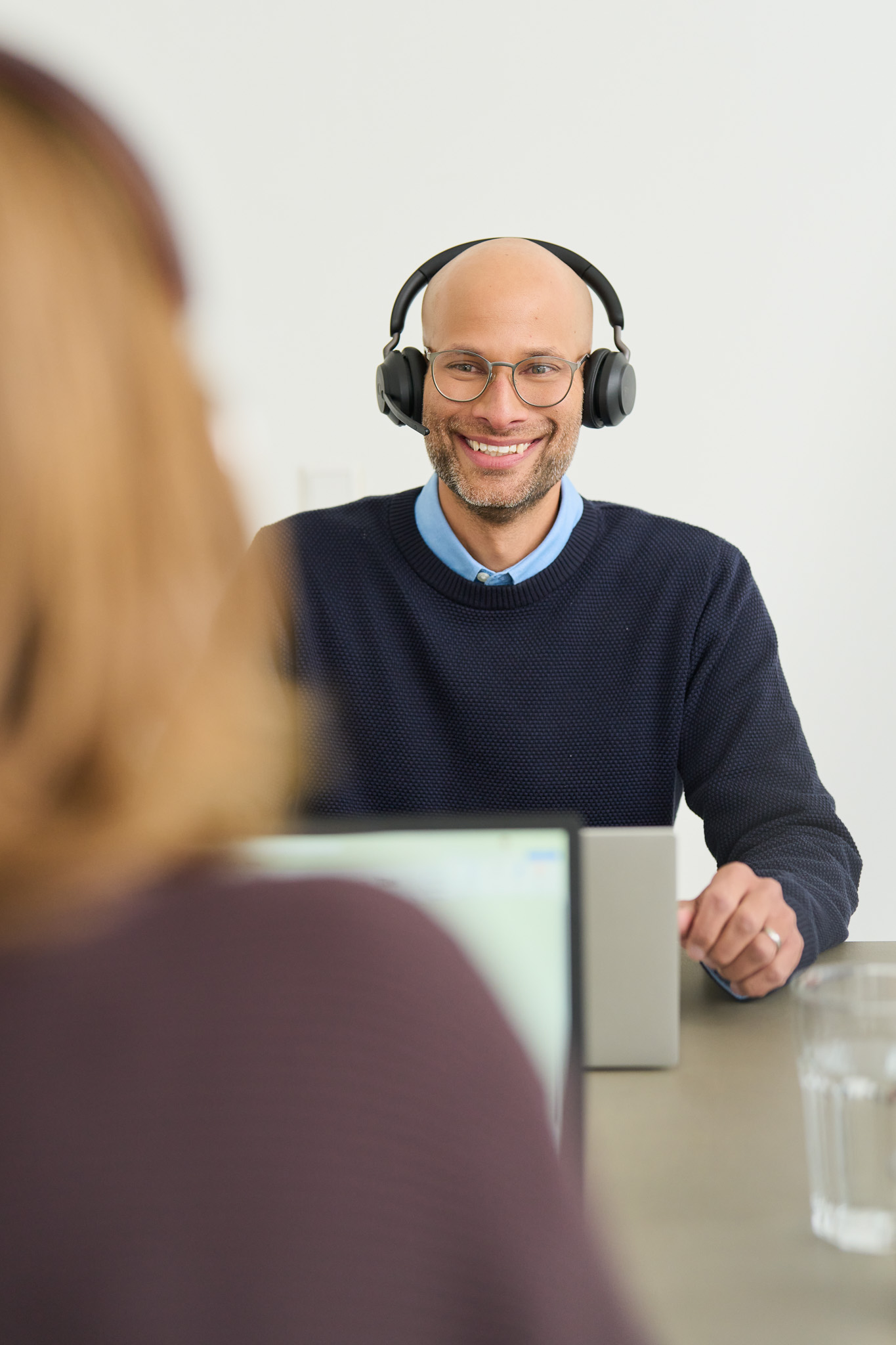 Ein Service-Mitarbeiter von loyos bi sitzt am Schreibtisch mit einem Laptop vor sich, er führt ein Gespräch mit Headset. Im Vordergrund der Rücken seiner Kollegin.