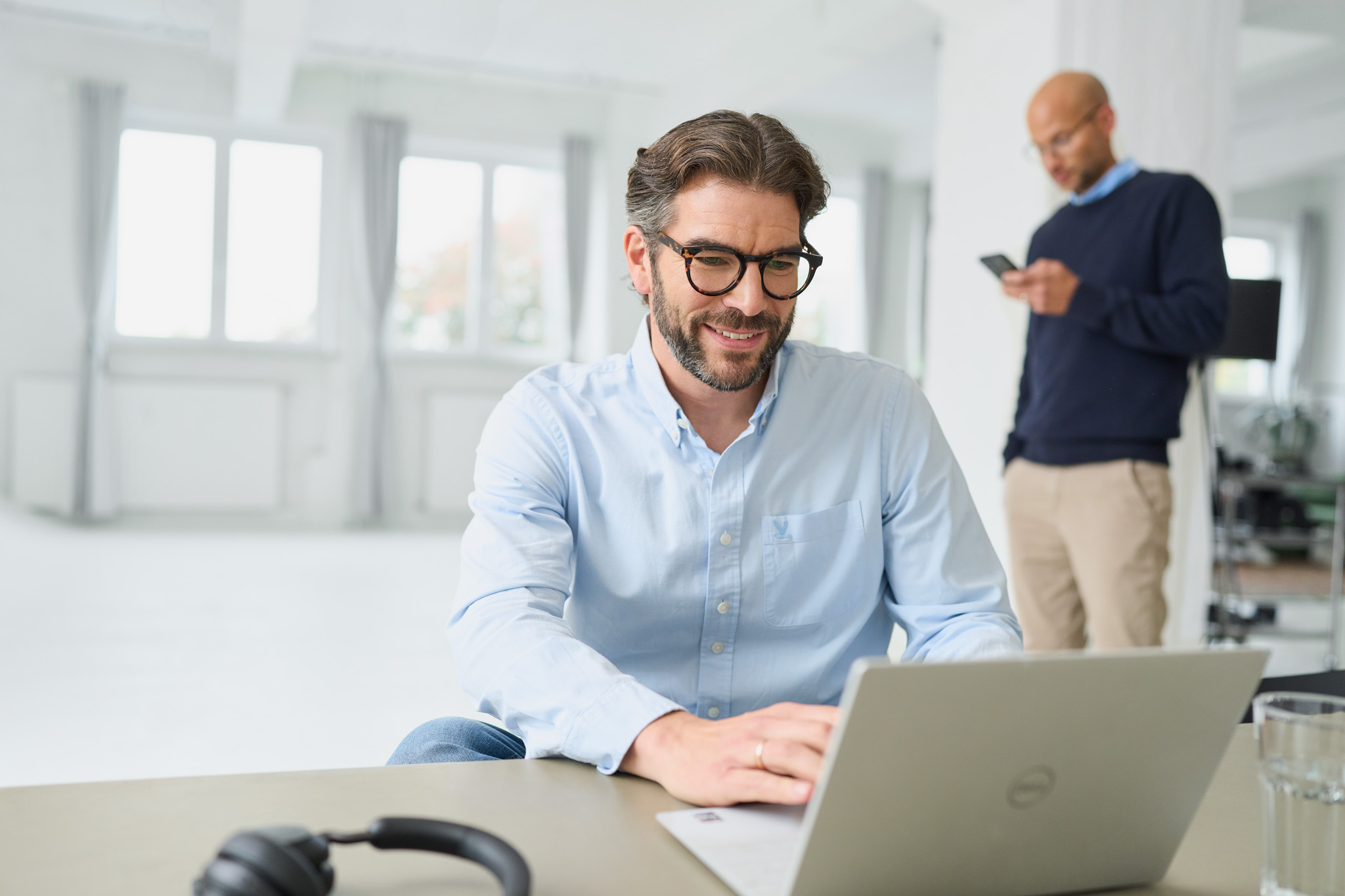 Martin Sperling von loyos bi lächelt in seinen Laptop an, ein Kollege mit Handy im Hintergrund