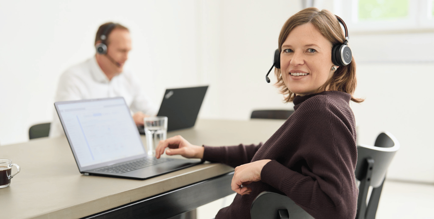 Eine Mitarbeiterin von loyos bi lehnt sich über den Stuhl, schaut direkt in die Kamera, vor sich der Laptop, im Hintergrund ein Kollege am Laptop. SIe tragen Headsets.