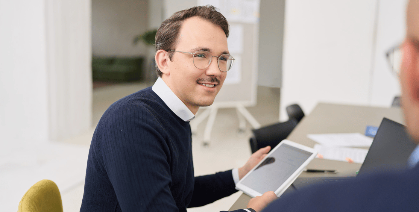 Florian von loyos bi sitzt am Schreibtisch, hat ein Tablet in der Hand und schaut seinen Kollegen an, den man nur angeschnitten von hinten sieht.