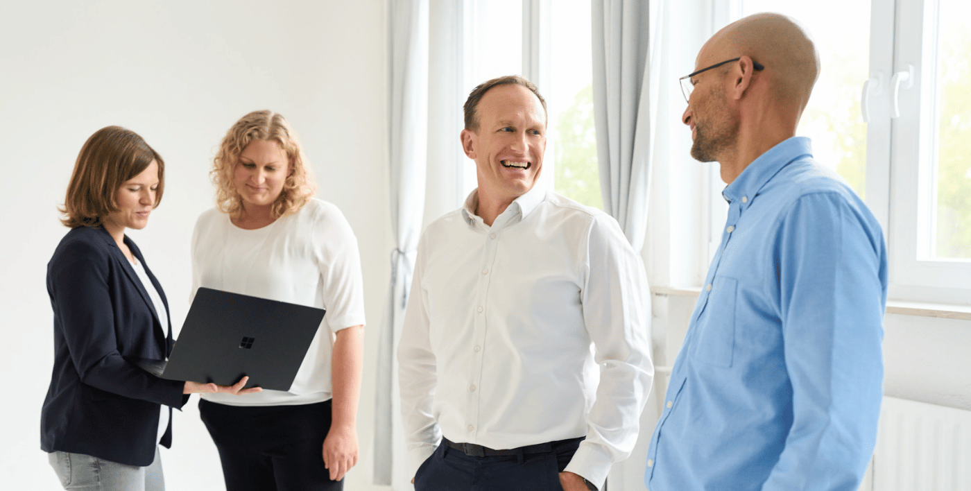 Zwei Männer im Vordergrund stehen und lachen sich an. Zwei Frauen im Hintergrund stehen auch und schauen auf einen Laptop. Sie alle arbeiten bei loyos bi.