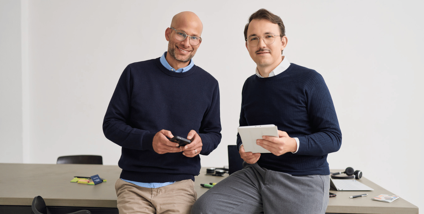 Zwei Mitarbeiter von loyos bi sitzen angelehnt an einen Tisch, lächeln in die Kamera. Sie tragen beide blaue Pullover.